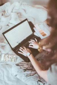 Fashion designer working on a laptop composing an email, with a phone showing a photo gallery and a color swatch book nearby, representing efficient design and tech pack work from home.