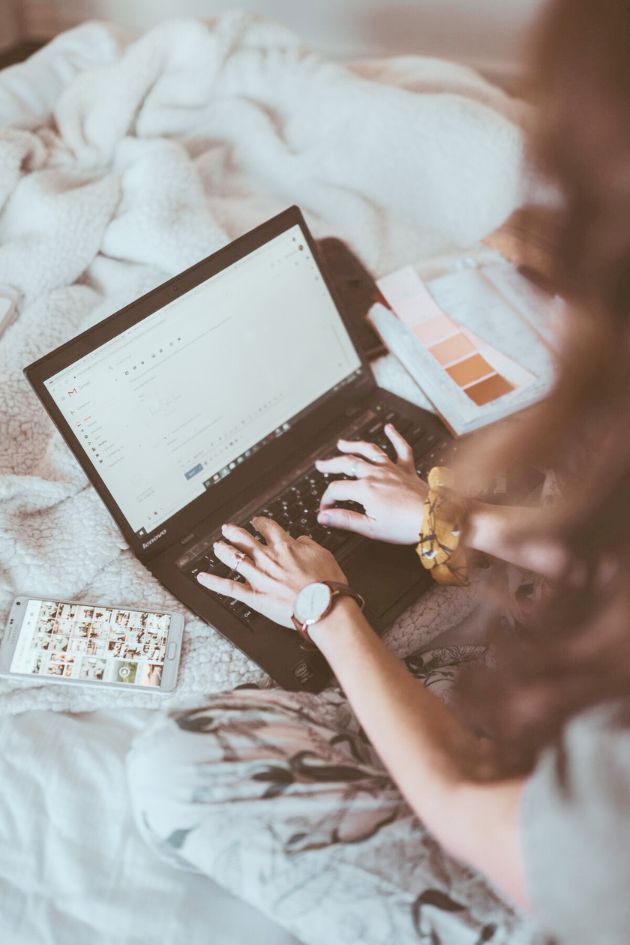 Fashion designer working on a laptop composing an email, with a phone showing a photo gallery and a color swatch book nearby, representing efficient design and tech pack work from home.