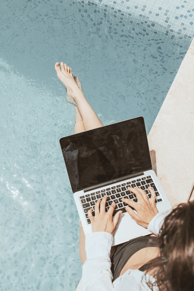 Overhead view of a person typing on a laptop with their feet dangling in a swimming pool.