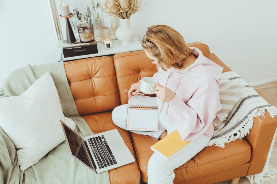 Woman sitting on a brown couch with a laptop, writing in a planner/notebook and holding a mug.