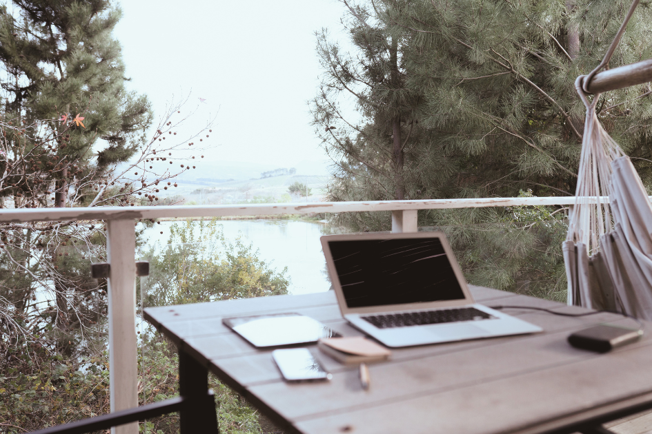 Laptop, tablet, and smartphone set up on a wooden table on a rustic outdoor balcony overlooking a scenic landscape.