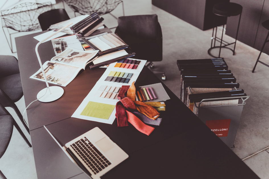 A fashion designer's workspace showing a desk with fabric swatches, a color card and laptop open
