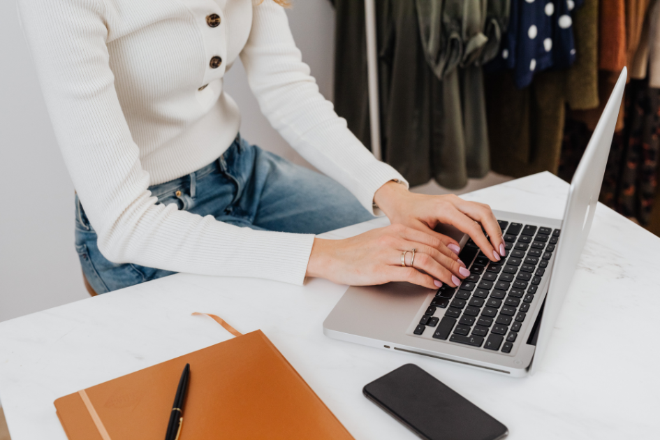 Woman working on laptop with clothes rack in background and notebook on desk beside computer
